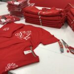 Pile di magliette e felpe con logo Sel Pulizie su tavolo con taglierini e forbice.